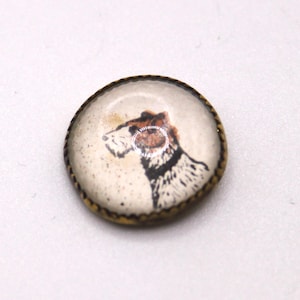 Puede incluir: Un broche redondo de estilo vintage con una ilustración de perro. El perro se representa de perfil con marcas marrones y negras sobre un fondo crema. El broche tiene un marco de metal dorado con un borde texturizado.