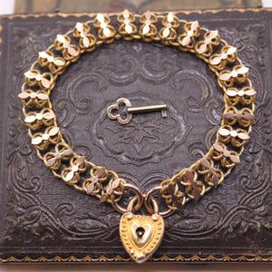 May include: A gold-toned bracelet with a heart-shaped lock and key, resting on a dark, textured box. The bracelet features a delicate floral design. The key is small and detailed. The box has an embossed pattern and gold trim.