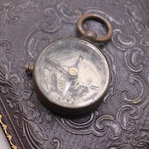 May include: Antique compass with a brass ring and a glass face, resting on a dark, textured surface with ornate embossed patterns. The compass needle points north. The surface has a gold trim.