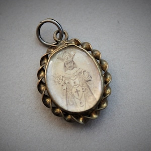 Op de afbeelding: Een ovale hanger in goudkleur met een glazen inzetstuk met een zwart-wit afbeelding van een persoon met een kroon en een gewaad. De hanger heeft een gedraaid touwontwerp rond de rand.