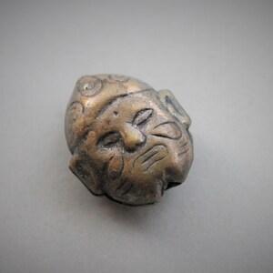 May include: A small, detailed, bronze-colored metal sculpture of a face with a textured surface.