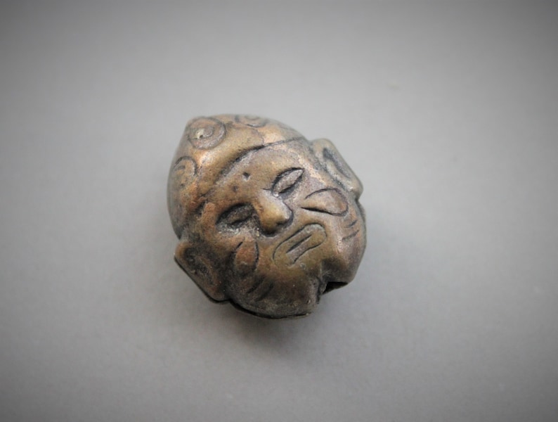 May include: A small, detailed, bronze-colored metal sculpture of a face with a textured surface.