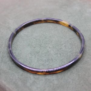 Può includere: Un braccialetto tondo con motivo tartarugato. Il braccialetto presenta una combinazione di colori marrone scuro e ambra. Il gioiello è realizzato in un materiale liscio e lucido ed è posto su uno sfondo verde tenue.