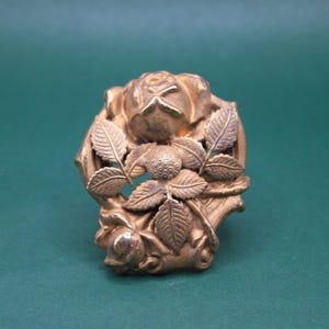 May include: A gold-colored, floral-themed brooch. The brooch features a detailed rose design with leaves and a bud. The metal has a slightly aged appearance, suggesting a vintage or antique piece. The brooch is set against a green background.