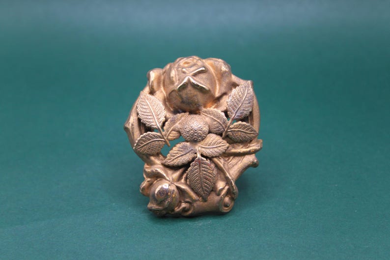 May include: A gold-colored, floral-themed brooch. The brooch features a detailed rose design with leaves and a bud. The metal has a slightly aged appearance, suggesting a vintage or antique piece. The brooch is set against a green background.