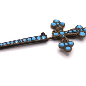May include: Vintage cross brooch with turquoise-coloured stones set in a bronze-toned metal. The brooch has a pin and a decorative bar with matching stones. The cross design is detailed and ornate.