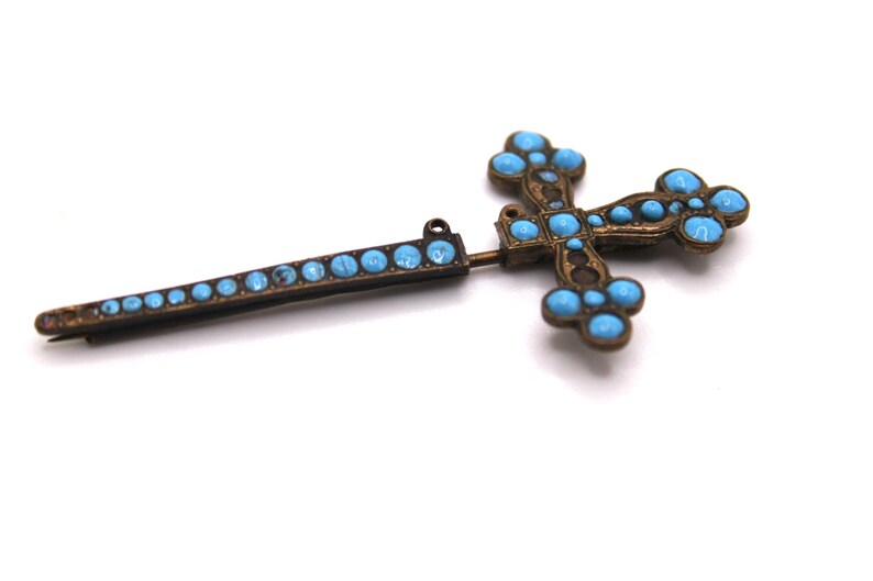 May include: Vintage cross brooch with turquoise-coloured stones set in a bronze-toned metal. The brooch has a pin and a decorative bar with matching stones. The cross design is detailed and ornate.
