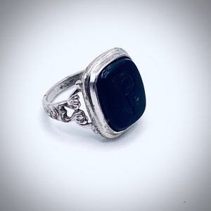 May include: A silver ring with a black onyx stone set in a square bezel. The stone has a carved letter 'P' on it. The ring has a delicate, ornate design.