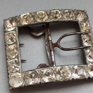 May include: A silver-toned buckle with a rectangular frame and a metal clasp. The frame is decorated with a row of clear glass stones.