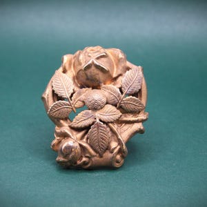 May include: A detailed, antique-style, gold-colored metal brooch. The brooch features a rose and leaf design. The rose is in the center, surrounded by detailed leaves. The background is a solid, dark green color.