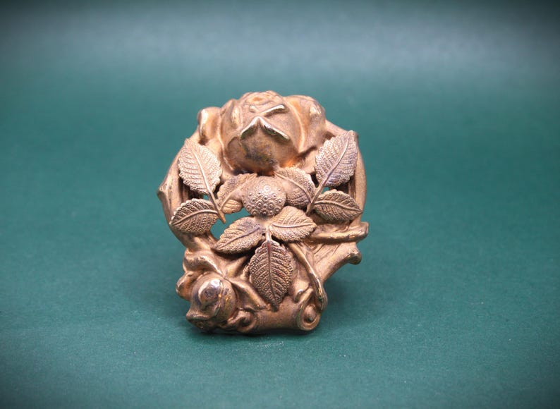 May include: A detailed, antique-style, gold-colored metal brooch. The brooch features a rose and leaf design. The rose is in the center, surrounded by detailed leaves. The background is a solid, dark green color.