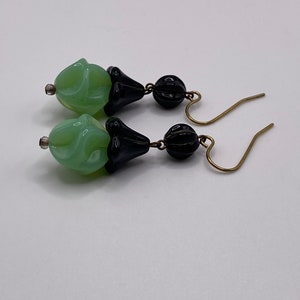 May include: A pair of dangle earrings with green and black glass beads. The beads are shaped like a flower and have a textured surface. The earrings have gold-toned hooks.
