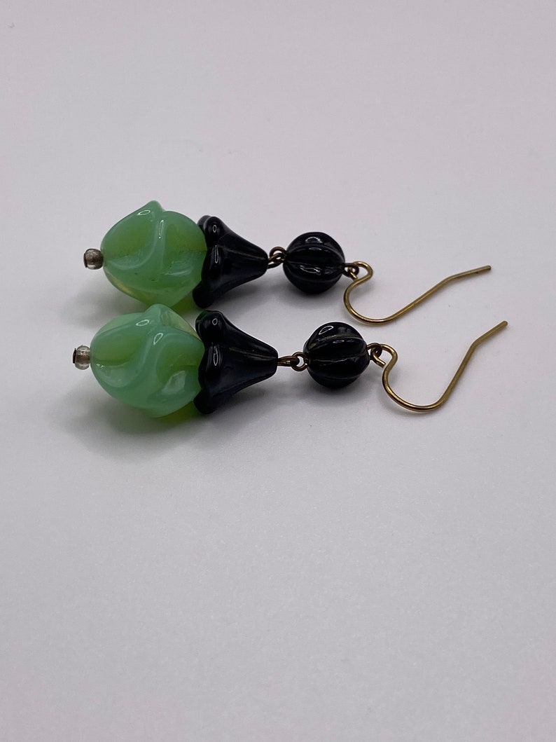 May include: A pair of dangle earrings with green and black glass beads. The beads are shaped like a flower and have a textured surface. The earrings have gold-toned hooks.
