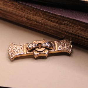 May include: A gold-toned antique brooch with intricate floral and scrollwork designs. The brooch features a central bow-shaped element with black and white accents. The piece is set against a neutral background, suggesting a vintage jewelry theme.