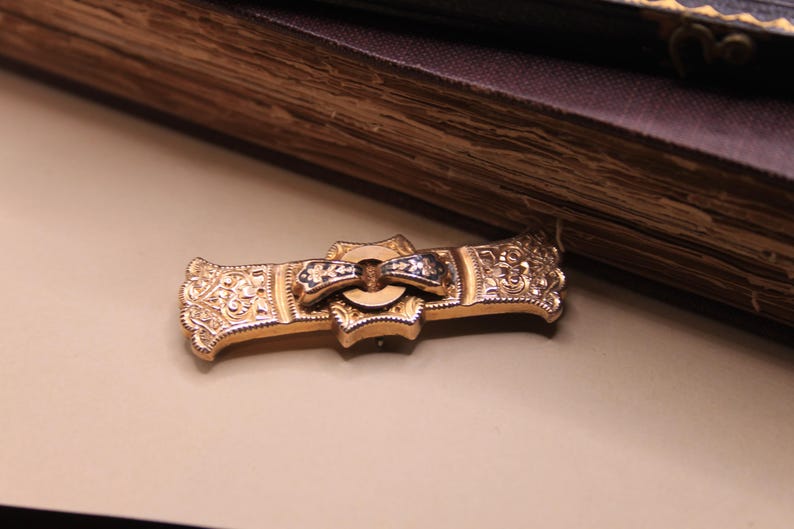 May include: A gold-toned antique brooch with intricate floral and scrollwork designs. The brooch features a central bow-shaped element with black and white accents. The piece is set against a neutral background, suggesting a vintage jewelry theme.