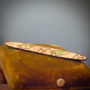 May include: A decorative enamel pin with a floral design in shades of pink, green, and white. The pin is resting on a brown velvet box.