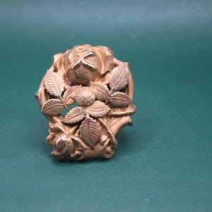 May include: A gold-colored, vintage-style brooch featuring a rose and leaf design. The brooch has a detailed, three-dimensional appearance, with the rose at the center and leaves surrounding it. The metal has a slightly aged look.