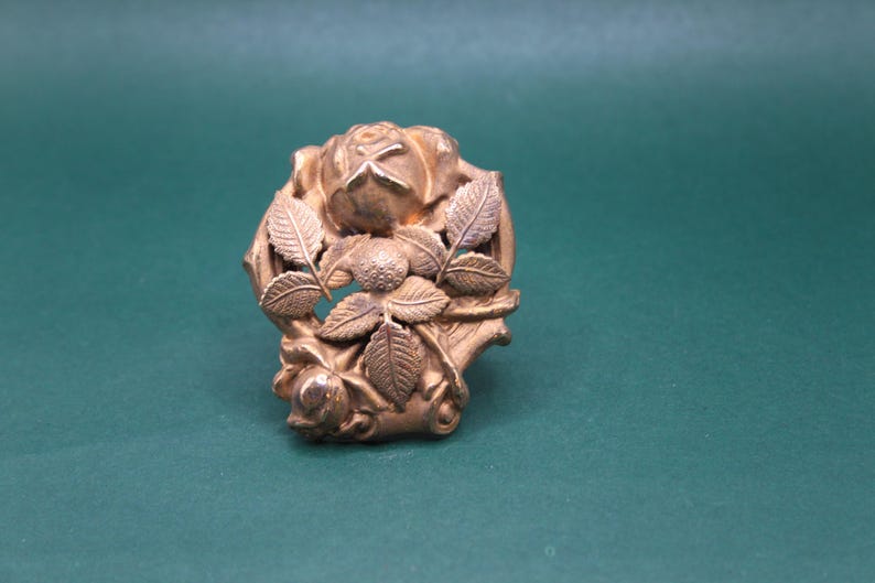 May include: A gold-colored, vintage-style brooch featuring a rose and leaf design. The brooch has a detailed, three-dimensional appearance, with the rose at the center and leaves surrounding it. The metal has a slightly aged look.