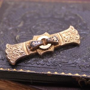 May include: Ornate gold-tone brooch with intricate floral and scrollwork designs. The brooch features a central buckle-like element with black enamel accents. It rests on a dark, textured surface with embossed patterns, suggesting a vintage jewelry piece.