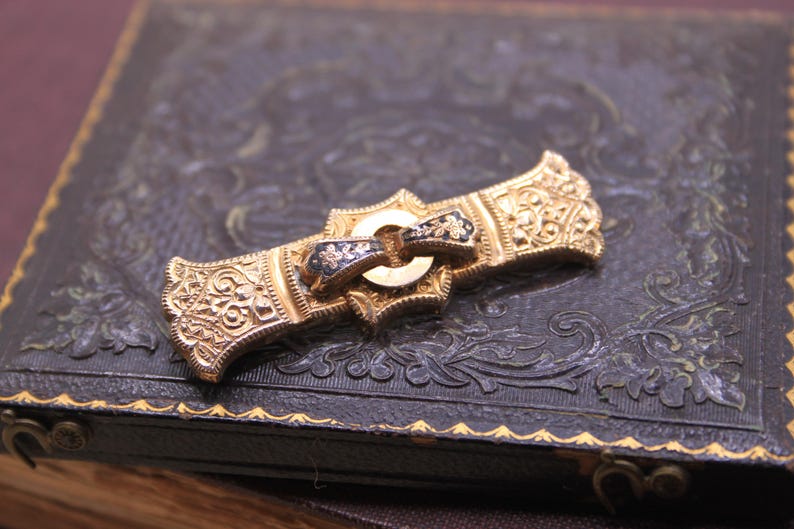 May include: Ornate gold-tone brooch with intricate floral and scrollwork designs. The brooch features a central buckle-like element with black enamel accents. It rests on a dark, textured surface with embossed patterns, suggesting a vintage jewelry piece.