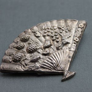 May include: A silver-toned brooch in the shape of a fan with intricate details. The fan is decorated with raised floral and leaf designs, and the center of the fan features a raised design of a woman's face.