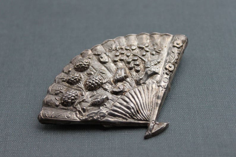 May include: A silver-toned brooch in the shape of a fan with intricate details. The fan is decorated with raised floral and leaf designs, and the center of the fan features a raised design of a woman's face.