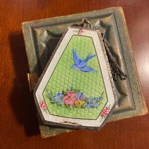 May include: A vintage silver-toned locket with a green background and a blue bird painted on the front. The locket is decorated with flowers and is resting on a green leather case.