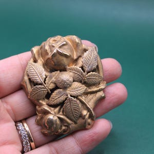 May include: A vintage gold-toned brooch featuring a detailed rose design with leaves and a central berry cluster. The brooch is held in a hand, showcasing its intricate floral details and aged patina.