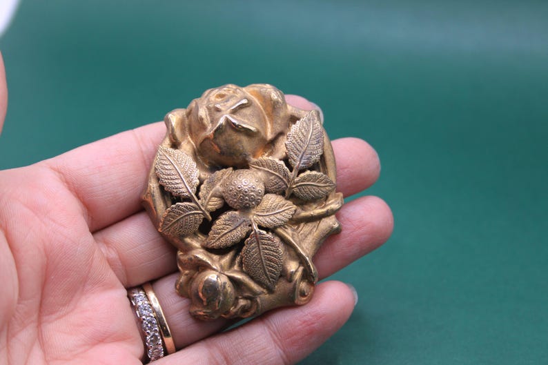 May include: A vintage gold-toned brooch featuring a detailed rose design with leaves and a central berry cluster. The brooch is held in a hand, showcasing its intricate floral details and aged patina.