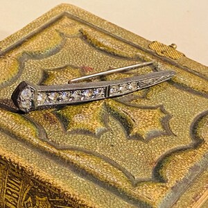 May include: A silver-toned metal brooch shaped like a railway spike, featuring a row of clear, faceted stones embedded along the length of the shaft and a larger stone at the head, with a thin metal pin mechanism attached to the back.