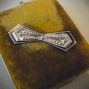 Peut inclure: Une broche en forme de n&oelig;ud en argent avec un motif g&eacute;om&eacute;trique, plac&eacute;e dans un &eacute;crin en velours.