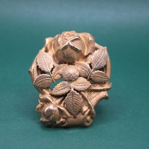 May include: A detailed, antique-style gold-colored metal brooch. The brooch features a central rose with surrounding leaves and smaller rose buds. The design is intricate and ornate, with a vintage aesthetic.