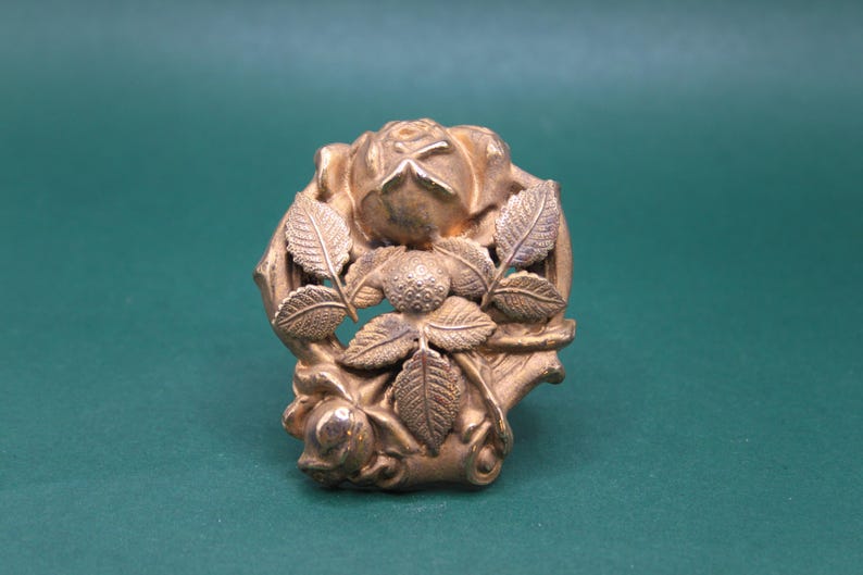 May include: A detailed, antique-style gold-colored metal brooch. The brooch features a central rose with surrounding leaves and smaller rose buds. The design is intricate and ornate, with a vintage aesthetic.