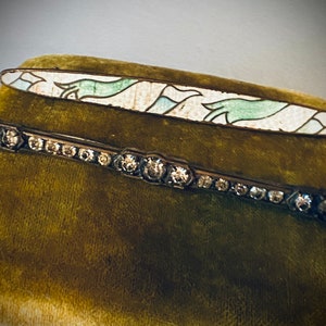 May include: Two vintage brooches on a green velvet background. The top brooch is a long, oval-shaped piece with a white enamel base and green and blue enamel designs. The bottom brooch is a silver-toned bar with a row of clear rhinestones.