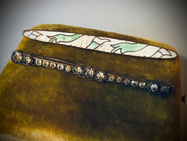 May include: Two vintage brooches on a green velvet background. The top brooch is a long, oval-shaped piece with a white enamel base and green and blue enamel designs. The bottom brooch is a silver-toned bar with a row of clear rhinestones.