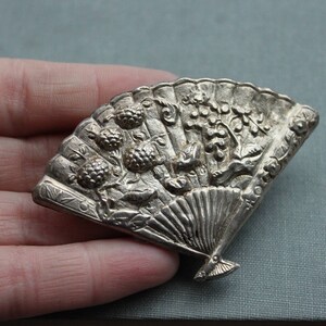May include: A silver-toned brooch in the shape of a fan with intricate floral and leaf designs. The fan is closed and has a detailed handle.