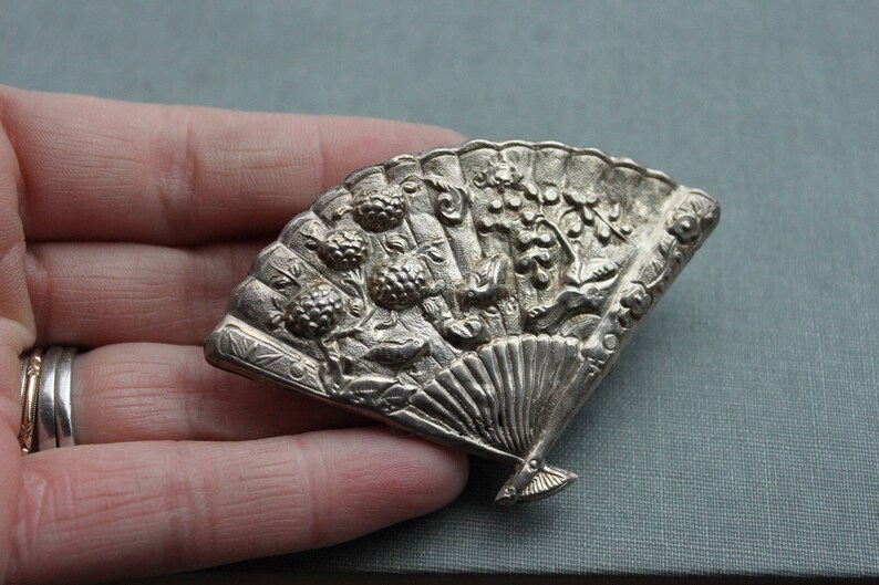 May include: A silver-toned brooch in the shape of a fan with intricate floral and leaf designs. The fan is closed and has a detailed handle.