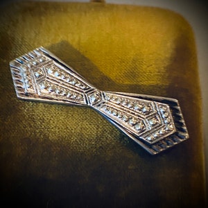 Peut inclure: Une broche en forme de n&oelig;ud papillon argent&eacute; avec un motif g&eacute;om&eacute;trique. La broche est pos&eacute;e sur un fond de velours vert.