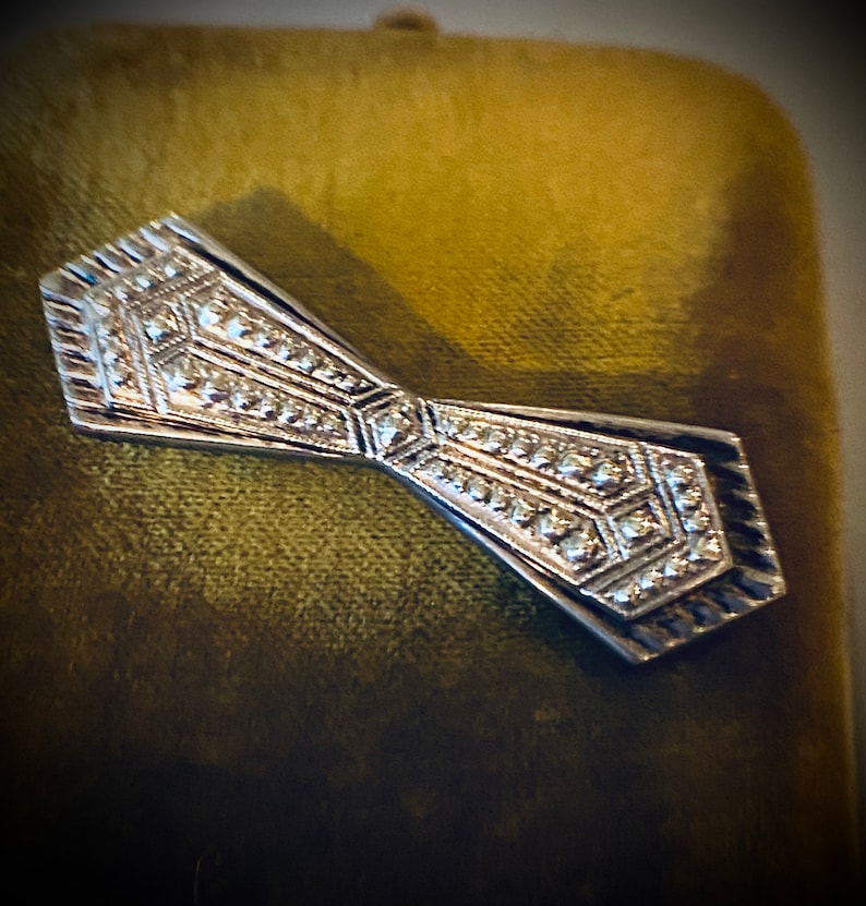 Peut inclure: Une broche en forme de n&oelig;ud papillon argent&eacute; avec un motif g&eacute;om&eacute;trique. La broche est pos&eacute;e sur un fond de velours vert.