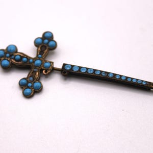 May include: A vintage cross brooch featuring turquoise-coloured stones set in a bronze-toned metal frame. The cross design is ornate. A straight pin, also with turquoise stones, extends from the cross.