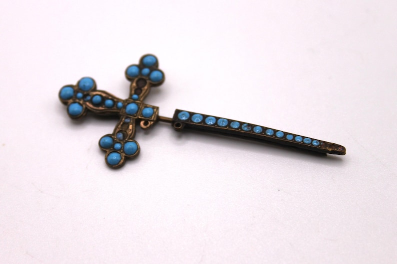 May include: A vintage cross brooch featuring turquoise-coloured stones set in a bronze-toned metal frame. The cross design is ornate. A straight pin, also with turquoise stones, extends from the cross.