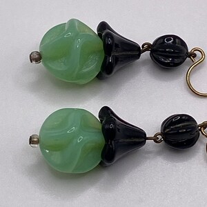 May include: A pair of dangle earrings with green and black glass beads. The green beads are round and have a swirl pattern. The black beads are shaped like a flower.