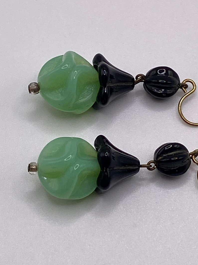 May include: A pair of dangle earrings with green and black glass beads. The green beads are round and have a swirl pattern. The black beads are shaped like a flower.