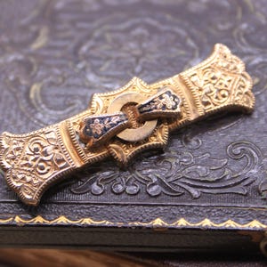 May include: Ornate gold-toned brooch with intricate filigree detailing. The brooch features a central clasp with blue and gold accents. The design is symmetrical and appears to be vintage or antique, with a detailed, textured surface.