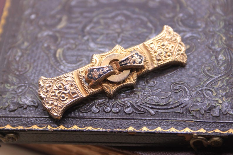 May include: Ornate gold-toned brooch with intricate filigree detailing. The brooch features a central clasp with blue and gold accents. The design is symmetrical and appears to be vintage or antique, with a detailed, textured surface.