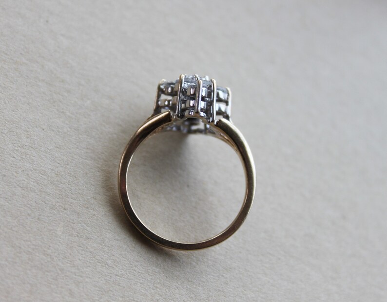 May include: A gold band ring with a large, clear, multi-faceted stone set in the center. The stone is surrounded by smaller, clear stones.