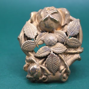 May include: A detailed, antique-style metal rose and leaf decorative element. The piece features a central rose bloom surrounded by detailed leaves, all in a tarnished gold color. The design is intricate and ornate.