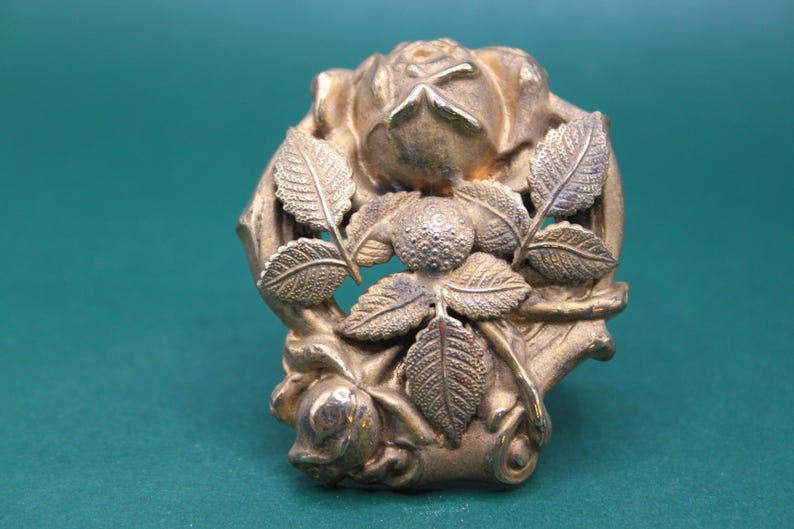 May include: A detailed, antique-style metal rose and leaf decorative element. The piece features a central rose bloom surrounded by detailed leaves, all in a tarnished gold color. The design is intricate and ornate.