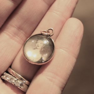 Puede incluir: Un colgante dorado con un pequeño retrato redondo en blanco y negro de una mujer en su interior. El colgante se sostiene en una mano, y se ven dos anillos en un dedo.