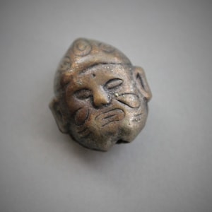 May include: A small, detailed, bronze-colored figurine of a man's face with a patterned hat. The figurine has a weathered look and appears to be made of metal.